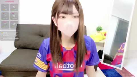 Video of a young Asian woman with long brown hair and a white mask, wearing a red and blue FC Barcelona soccer jersey, seated on a dark brown couch in a minimalistic room with a calendar and a plant in the background.