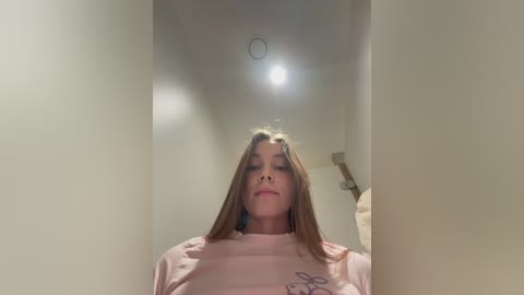 Video of a young woman with straight, light brown hair, wearing a light pink t-shirt, standing in a narrow, beige hallway with a bright ceiling light above.