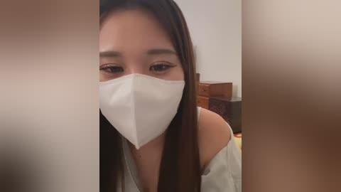 Video of a young Asian woman with long, straight brown hair, wearing a white surgical mask and a white off-shoulder top, looking directly at the camera with a neutral expression.