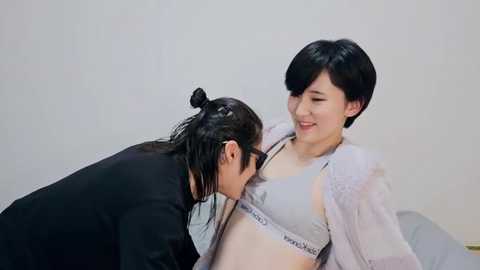 Video of a smiling pregnant woman in a grey Calvin Klein sports bra, being kissed by a woman with black hair in a ponytail, wearing a black top. Both in a simple, light-colored room.