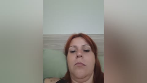 Video of a plus-sized, fair-skinned woman with shoulder-length, red hair and makeup, looking downward. She wears a black top and is framed by a beige wall with white molding. The image is slightly out of focus.