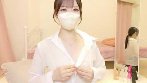 A video of an Asian woman with fair skin, dark hair, and a white face mask, wearing a white unbuttoned shirt, standing in a pink room with a bed and mirror, holding a bottle.
