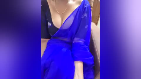 Video of a light-skinned woman wearing a sheer, blue sari draped over a black, low-cut blouse, revealing cleavage. The background is blurred, suggesting an indoor setting.