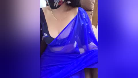 A video of a woman wearing a sheer blue sari draped over a black bra, revealing her medium-sized breasts, sitting indoors with a blurred background.