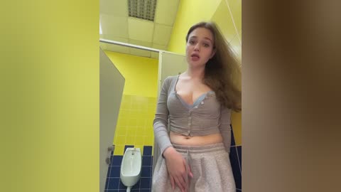 A video of a young Caucasian woman with long, light brown hair, wearing a grey crop top and skirt, standing in a yellow-tiled bathroom with blue-tiled floor and a urinal.