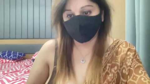 A video shows a light-skinned woman with long brown hair, wearing a black face mask, beige top, and gold necklace, sitting on a bed with patterned sheets.