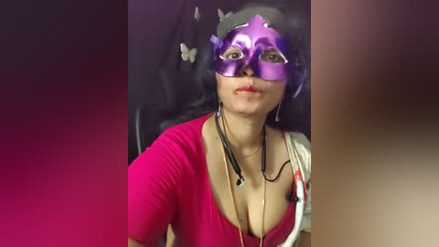 Video of a woman in a purple Venetian mask, red top, and gold necklace, seated with a blurred background, emphasizing her fair skin and subtle makeup.