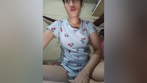 A video of a young woman with medium skin tone, wearing a light blue t-shirt with cherry print, sitting on a bed with a white sheet, in a dimly lit room.