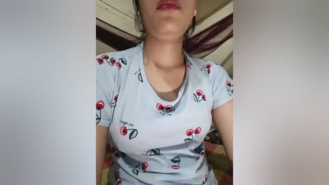 A video of a light-skinned woman with dark hair, wearing a white T-shirt with cherry print, partially cropped at the neck and face, in an indoor setting with visible wooden beams and a colorful blanket.