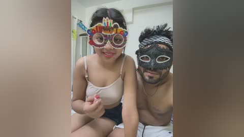 A video of an Asian woman with medium-sized breasts in a white tank top, wearing a colorful party mask, sitting next to a shirtless man in a black mask, in a bedroom.