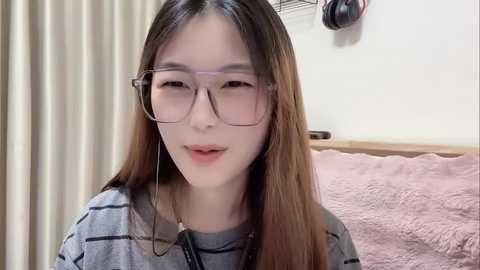 Video of an Asian woman with long, straight brown hair and clear glasses, wearing a gray striped shirt, smiling softly in a cozy bedroom with a pink quilted blanket and beige curtains.