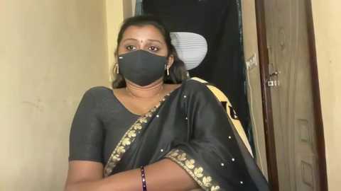 Video of an Indian woman in a black saree with gold embroidery, wearing a black mask and a black top, standing in a dimly lit doorway, arms crossed.