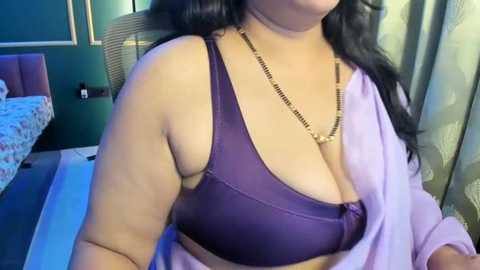 Video of a woman with long black hair, wearing a purple bra and a light pink robe, sitting in a modern bedroom with a bed and green curtains.