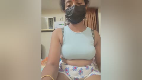 Media: Video of a woman with medium skin tone, wearing a black mask, light blue crop top, and floral shorts, sitting on a bed with beige walls and a window in the background.