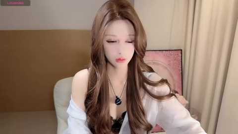 Video of a fair-skinned woman with long, wavy brown hair, wearing a white off-shoulder blouse and black lace bra, sitting pensively indoors.