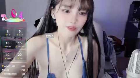 A video of an East Asian woman with long black hair, wearing a blue bra, sitting on a bed in a dimly lit room.