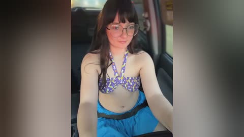 Video of a young woman with long dark hair, fair skin, and glasses, wearing a purple and yellow patterned bikini top and blue shorts, sitting in a car.