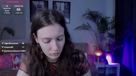 A video of a young woman with long brown hair, wearing a floral blouse, sitting indoors. The background features a TV, potted plants, and framed pictures. A livestream overlay with viewers and comments is visible.
