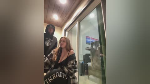 Video of a dimly lit hallway featuring two women, one with long hair in a black hoodie, the other with a plaid shirt, both with a casual demeanor.