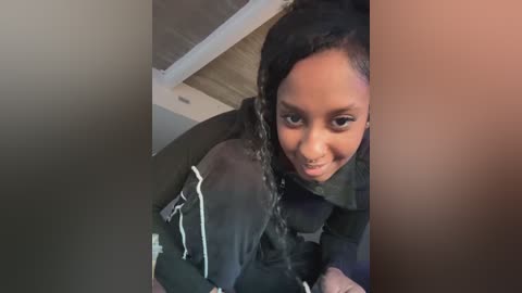 Video of a young Black girl with braided hair, wearing a black jacket and a scarf, smiling, with a blurred background.