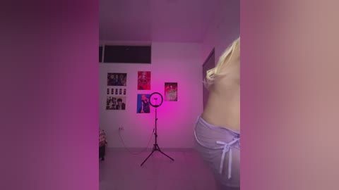 Video of a dimly lit, sparsely decorated room with a large round light on a stand, purple walls, and a partially visible person in light blue pajama pants.