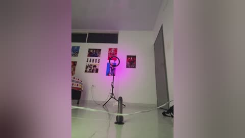 Video of a dimly lit, sparsely furnished room with white walls, a single large window, and a small black stand with a ring light emitting purple light. The room appears to be in the process of being set up for videoy.