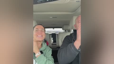 Video of a smiling woman in a green hoodie, with a bald man in a black jacket, seated in a car with beige seats.
