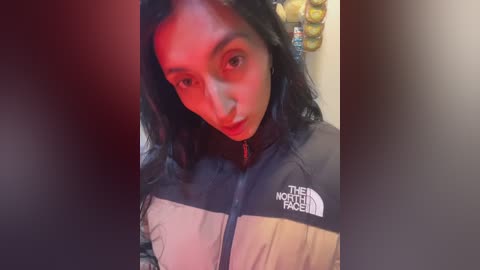 Video of a young woman with long black hair, wearing a The North Face jacket, standing in a dimly lit room with red lighting.