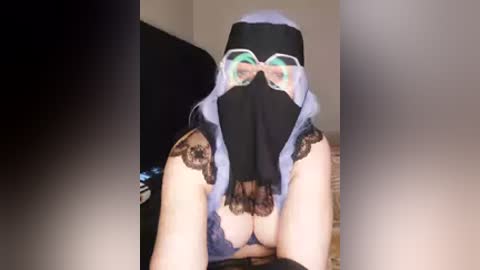 Video of a woman in a black nun's habit, with a lace-trimmed bra visible, revealing her breasts. She wears green-tinted goggles and her face is obscured by the habit's hood. Background is a dark room with indistinct furniture.
