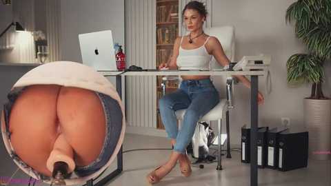 Video of a woman in a white tank top and jeans, seated at a desk with a laptop, a dildo in a glass display, and office supplies.