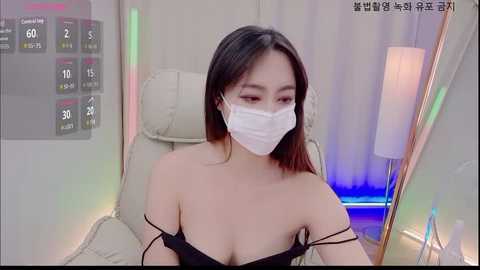 Media: Video of an Asian woman with long black hair and fair skin, wearing a white surgical mask and a black off-shoulder top, sitting in a beige armchair in a modern room with a colorful LED light strip.