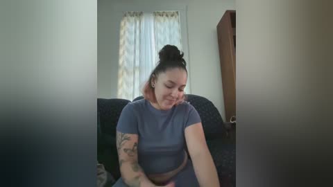 Video of a smiling, tattooed, light-skinned woman with dark hair in a bun, wearing a gray crop top, sitting on a dark sofa in a dimly lit room with light curtains.