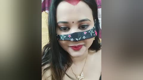 Video of a smiling South Asian woman with long black hair, wearing a black mask with red stars and flowers, and a gold necklace. She has light skin and red lipstick.