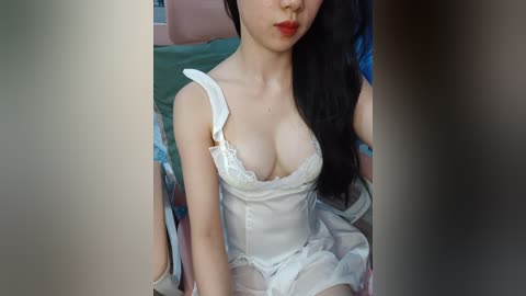 Video of an Asian woman with long black hair, wearing a white lace lingerie top, showing cleavage, sitting on a bed with green and blue sheets.
