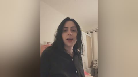 A video of a young, light-skinned woman with long black hair, wearing a black button-up shirt, standing in a dimly lit room with beige walls, a door, and a white curtain.