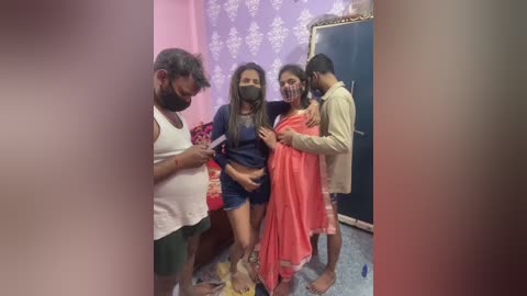 Video of four people in a small, dimly lit room with purple and white wallpaper. Two men and two women, all with brown skin tones, wearing casual clothes. One woman is masked.