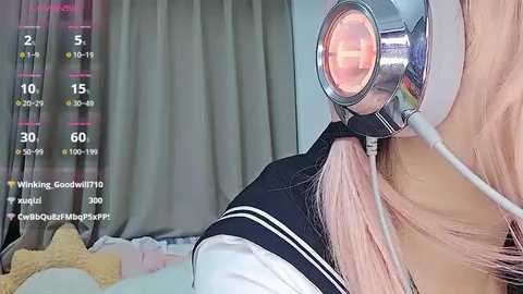 Video of a young woman with long pink hair, wearing a black and white schoolgirl uniform, with a futuristic, glowing headset. Background features a bed with a beige blanket, and a screen displaying game stats.