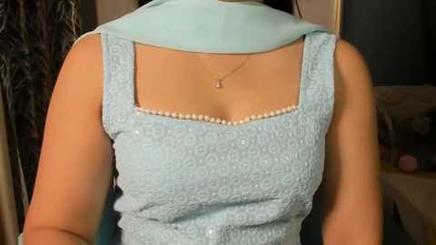 Video of a woman's upper torso wearing a light blue, textured top with a square neckline and a matching light blue scarf. She wears a delicate gold necklace. Background shows a dark curtain and a cluttered room.