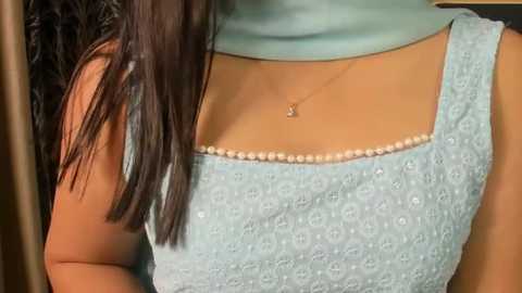 Video of a woman's upper body in a light blue embroidered kurta, showcasing a delicate necklace with a small pendant. Her long, straight black hair falls over her shoulders.