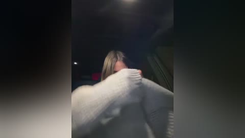 Media: A blurry, nighttime photo of a person, possibly male, with shoulder-length blonde hair, wearing a white sweater, hunched over, looking down. The background is dimly lit, suggesting a car interior.