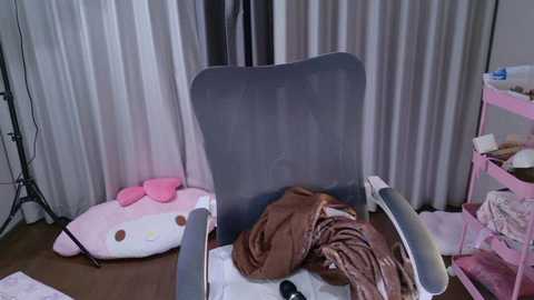 Video of a messy room with a gray recliner, brown clothes, a pink Hello Kitty pillow, and white curtains.