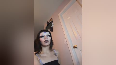Media: Video of a young woman with pale skin and dark hair, wearing a black dress, white face paint, and a headband. She stands in a hallway with a beige wall and a closed door.