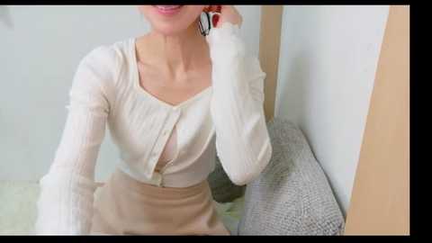 Video of a smiling woman with fair skin, wearing a cream-colored cardigan and beige skirt, seated on a grey armchair against a white wall.