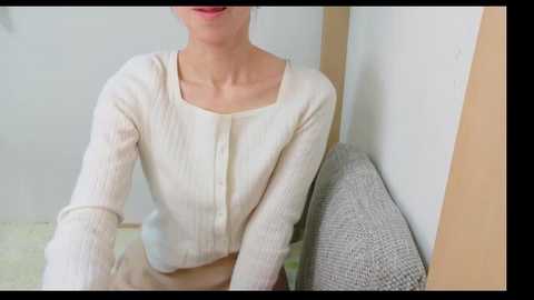 Video of a slender, light-skinned woman in a cream cardigan, seated on a gray textured chair. Background features white walls and a beige corner.