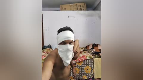 Video of a shirtless South Asian man with a white mask, sitting on a bed with a floral-patterned bedspread, in a cluttered room with cardboard boxes.