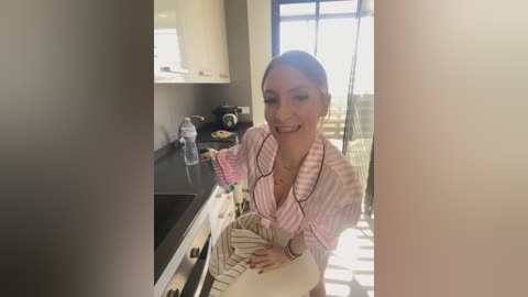 A video captures a smiling, light-skinned woman with brown hair in a ponytail, wearing a pink-striped robe, standing in a modern kitchen with white cabinets and a black countertop.