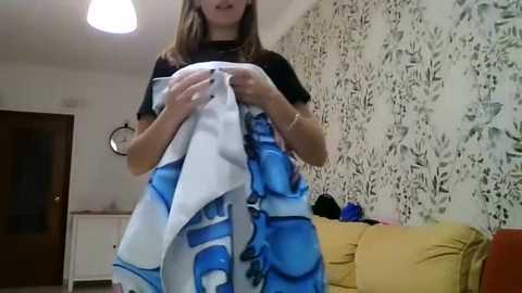 Video of a young woman with light brown hair, wearing a black t-shirt, holding a blue and white patterned towel, standing in a cozy living room with floral wallpaper and a mustard-yellow couch.