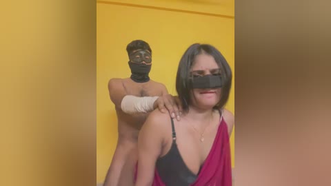 Video of a man with bandaged face and chest, blindfolded, standing behind a woman with dark hair, black bra, red shawl, and blindfold, against a yellow background.