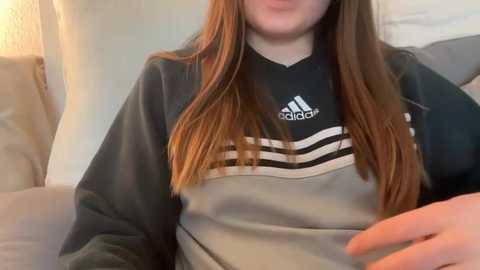 Video of a young woman with long, straight, auburn hair wearing a black Adidas sweatshirt with white stripes. She's sitting on a light-colored couch, her hand resting on her lap.