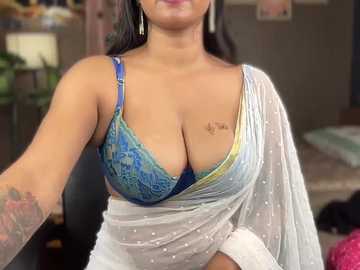 Video of a South Asian woman with medium brown skin and long black hair, wearing a blue and yellow bra and a white saree, with a tattoo on her right breast. Background shows a blurred room with furniture.